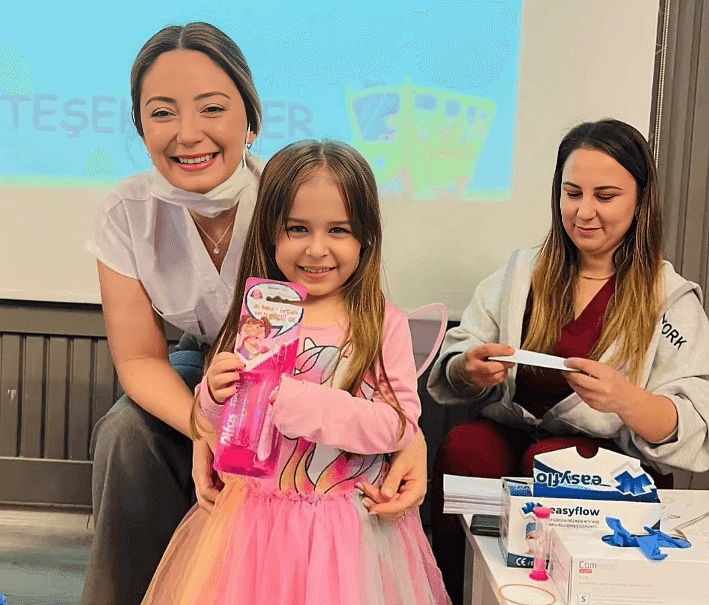 A smiling girl in a pink dress holding a gift, accompanied by two healthcare professionals