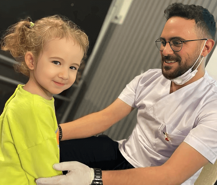 A smiling dentist in a white coat holding a young girl in a bright yellow shirt during a dental event